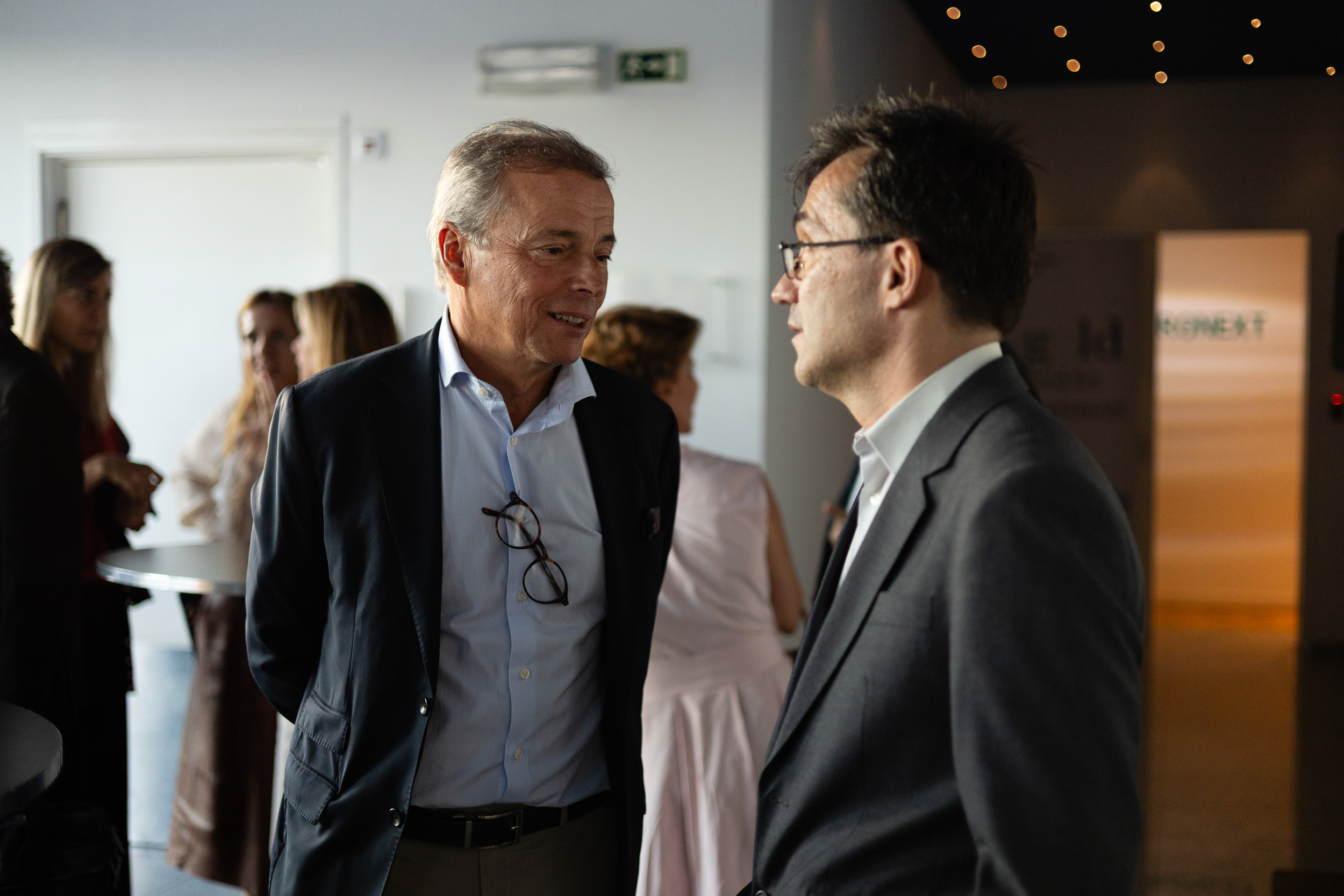Two men in suits converse at a social event, with glasses hanging from one man's shirt, in a softly lit room.