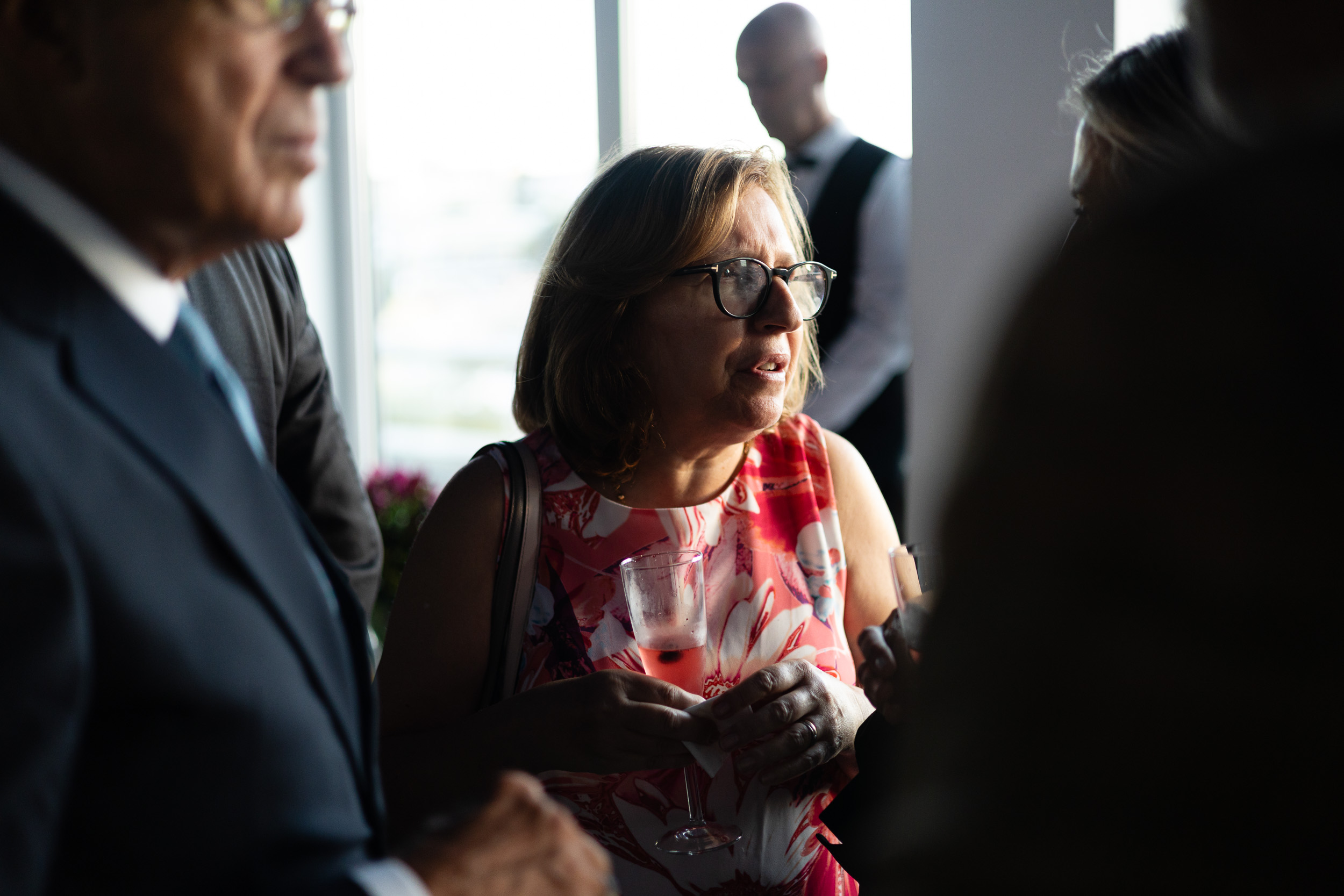 Center: woman with short hair and glasses in a pink floral dress, talking and holding a champagne flute at a social gathering in soft focus background.