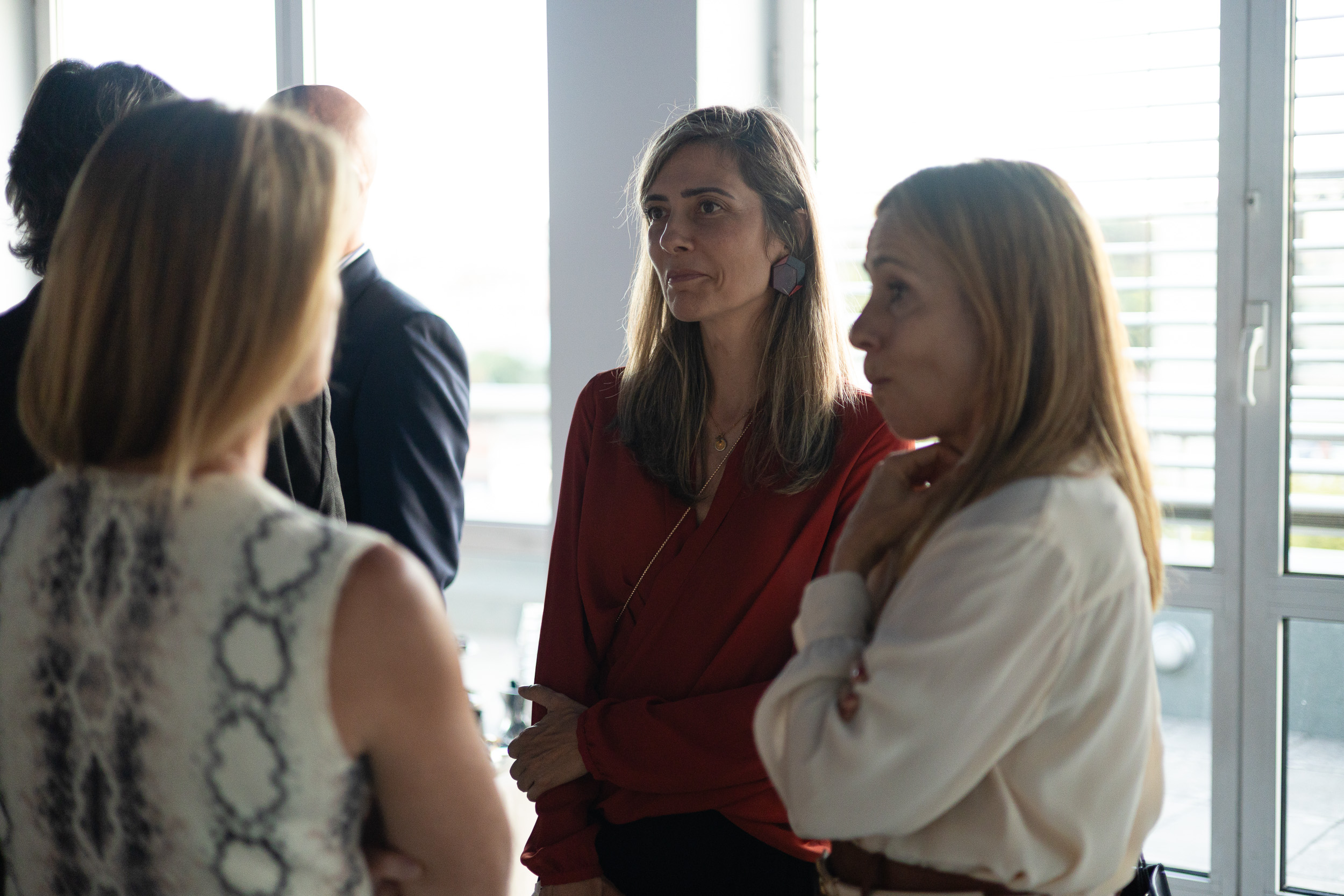 Woman in a red blouse speaks with colleagues in a bright room by large windows