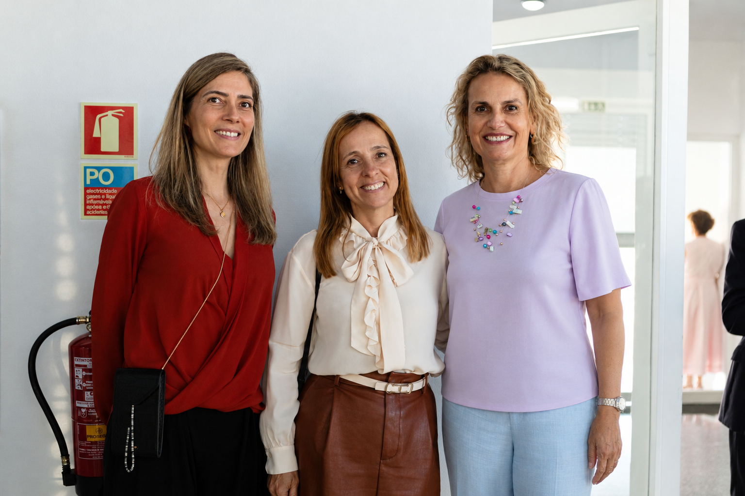 Three women smiling and posing together in a bright hallway, dressed in business casual.